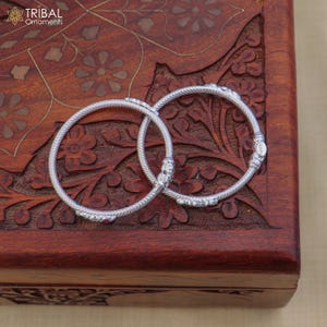 May include: A pair of silver baby bangles with a textured design and decorative details. The circular bangles are displayed on a dark wooden surface with carved floral patterns. The jewellery is from Tribal Ornaments.