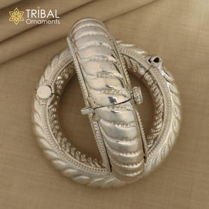 May include: Two silver bangles with intricate designs. The bangles are open and have a hinged clasp. The bangles are made of a silver metal and have a shiny finish.