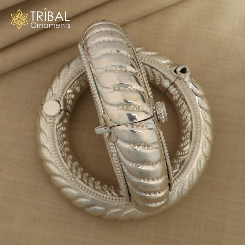 May include: Two silver bangles with intricate designs. The bangles are open and have a hinged clasp. The bangles are made of a silver metal and have a shiny finish.