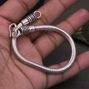 K&ouml;nnte beinhalten: Ein silbernes Schlangenkettenarmband mit einem Verschluss. Das Armband besteht aus einer d&uuml;nnen, flexiblen Kette, die wie eine Schlange aussieht.