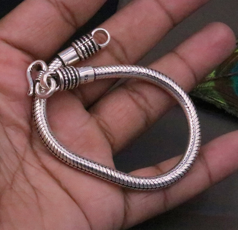 K&ouml;nnte beinhalten: Ein silbernes Schlangenkettenarmband mit einem Verschluss. Das Armband besteht aus einer d&uuml;nnen, flexiblen Kette, die wie eine Schlange aussieht.
