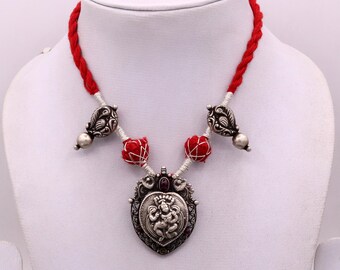 May include: A silver necklace with a red and white cord. The pendant is a heart-shaped design with a silver deity figure inside. The necklace has small silver beads and decorative silver charms.