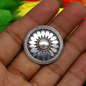 May include: A silver oval pendant with a floral design. The pendant features a central circle with a raised, textured surface surrounded by a ring of petals.