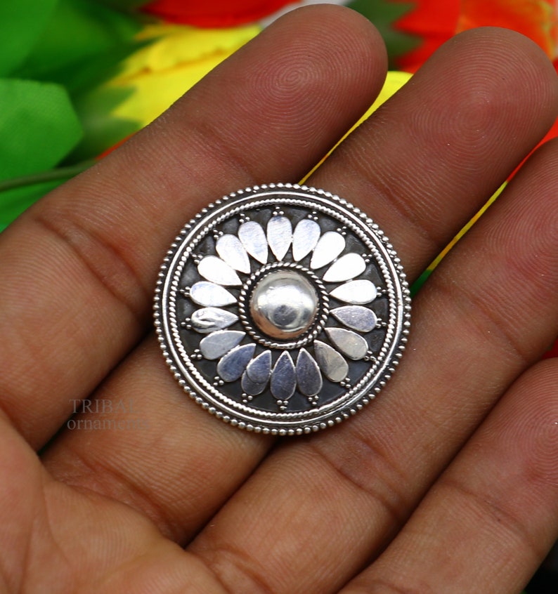 May include: A silver oval pendant with a floral design. The pendant features a central circle with a raised, textured surface surrounded by a ring of petals.