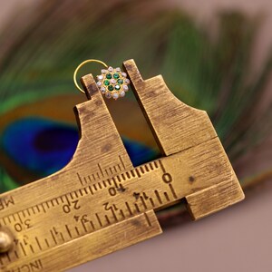 May include: A gold nose ring with a circular design featuring green and white gemstones. The ring is being measured with a brass caliper.