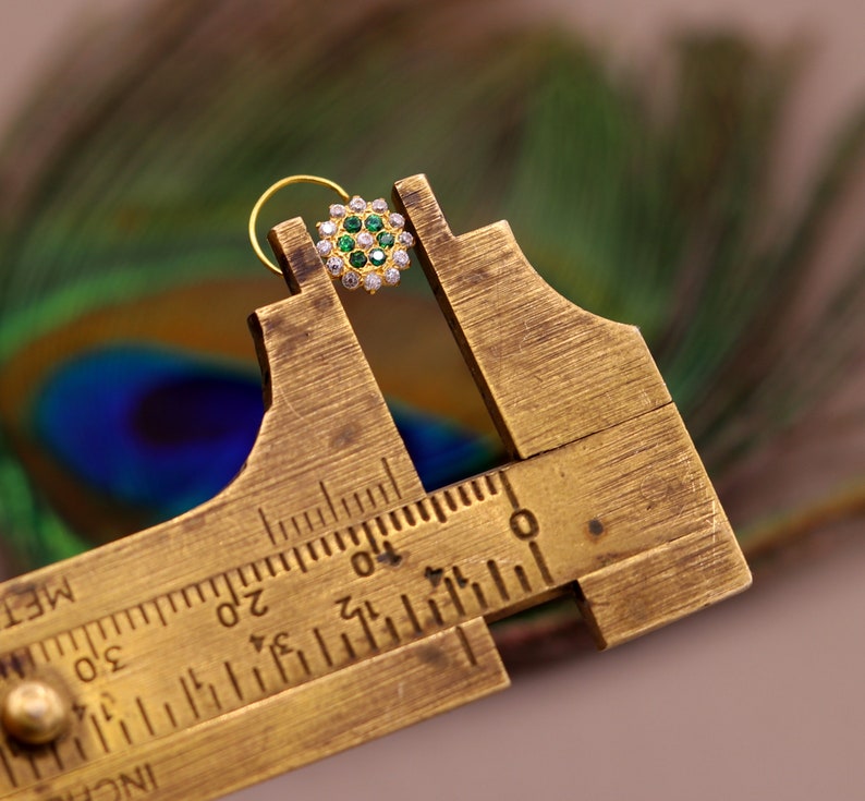 May include: A gold nose ring with a circular design featuring green and white gemstones. The ring is being measured with a brass caliper.