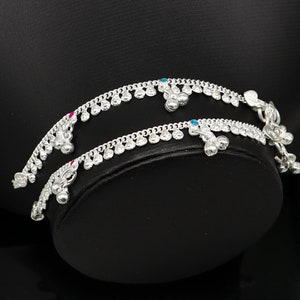 May include: Two silver anklets with small bells and blue and pink gemstones. The anklets are on a black surface.