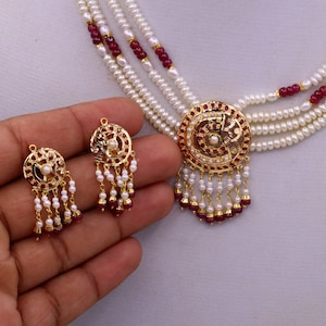 May include: A gold necklace with a round pendant featuring red stones and white pearls. The necklace is layered with four strands of white pearls and red stones. Matching earrings with a similar design are also included.