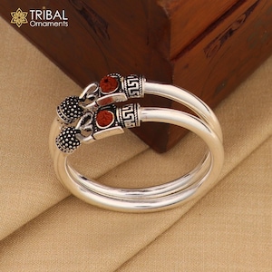 May include: A pair of silver anklets. Each anklet has a decorative section with a red stone and a patterned design. The anklets are displayed on a tan surface, with a wooden box in the background. The logo "TRIBAL Ornaments" is visible.
