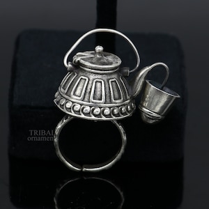 May include: A silver ring with a teapot design. The teapot has a small handle and a spout. There is a small silver bucket attached to the spout.