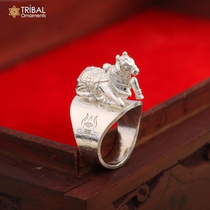 May include: A silver ring featuring a detailed sculpture of a cow. The ring has an open design with an engraved symbol on the band. The ring is displayed on a wooden surface with a red background. The Tribal Ornaments logo is in the top left corner.