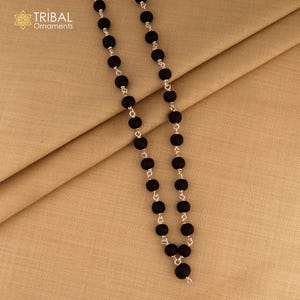 May include: A black beaded necklace with silver-toned links. The necklace features round, matte black beads strung together with small, silver-colored connectors. The necklace is displayed on a tan fabric background. The upper left corner has the text "TRIBAL Ornaments".
