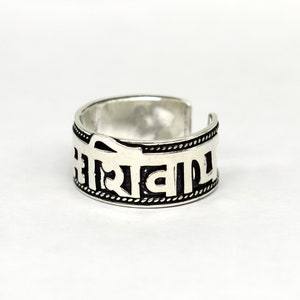 May include: A silver ring with a black inscription in a Sanskrit-like script. The ring is open ended and has a textured band.