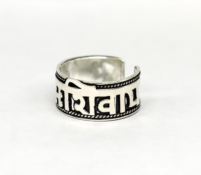 May include: A silver ring with a black inscription in a Sanskrit-like script. The ring is open ended and has a textured band.