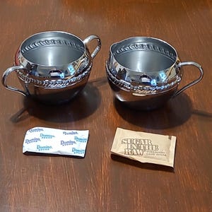 May include: Two silver sugar bowls with ornate detailing and a brown packet of "Sugar in the Raw" turbinado cane sugar.