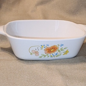 May include: White ceramic baking dish with a floral design featuring orange, yellow, and blue flowers. The dish has two handles on the sides.