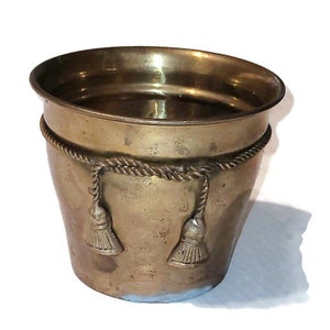 May include: A brass planter with a rope design around the rim and tassels hanging from the rope. The planter is a light gold color and has a slightly distressed look.
