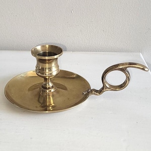 May include: A vintage brass candlestick holder with a circular base and a handle. The candle holder has a cup-shaped top and a slender stem. The handle is a loop shape. The brass has a warm, golden hue.