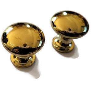May include: Two gold-toned cabinet knobs with a round top and a slightly flared base.