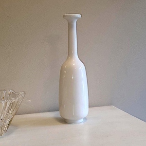 May include: A tall, white ceramic vase with a slender neck and a rounded body. The vase has a smooth, glossy finish and stands on a small, circular base. The design is minimalist and elegant, suitable for displaying flowers or as a decorative piece.