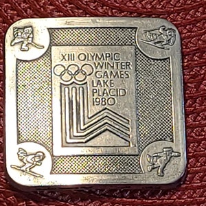 May include: A silver square pin commemorating the XIII Olympic Winter Games in Lake Placid 1980. The pin features the Olympic rings, the games' logo, and illustrations of winter sports. The pin is set against a textured, reddish-brown background.