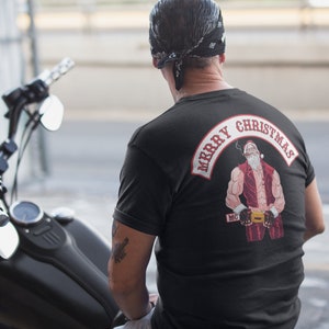 Op de afbeelding: Zwart T-shirt met een rood-witte "Merry Christmas" patch met een gespierde kerstman die een sigaar vasthoudt en een rood-witte riem draagt met "MC" erop.