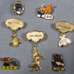 May include: A collection of seven gold-tone metal bowling achievement pins. The pins feature various designs including a 150 club shield, a cat with a bowling ball, 190, 180, and 160 club plaques with dangling charms, a 500 eagle, and a 500 bear.