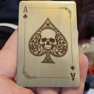 May include: A metallic Ace of Spades playing card featuring a skull design within the spade symbol. The card has decorative corner flourishes and the letter "A" with a small spade symbol in the corners.