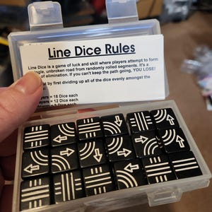 May include: A clear plastic case containing black Line Dice with white line and arrow designs. The case also holds a card with the game's rules. The dice are square and feature various line and arrow patterns.