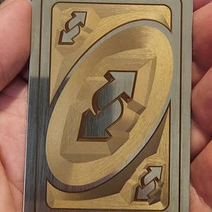 Puede incluir: Una tarjeta metálica con acabado dorado y plateado. La tarjeta presenta un diseño ovalado central con un símbolo de flecha invertida, rodeado por un borde plateado. Símbolos de flecha invertida adicionales están en cada esquina. La tarjeta se sostiene en una mano.