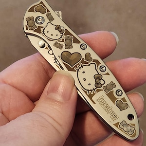 May include: A silver folding knife with a decorative design featuring hearts, bows, and the Hello Kitty character. The knife has the text "Kershaw Speedsafe USA" printed on the side.