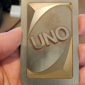 May include: A metal UNO card with a brushed silver border and a gold-toned center. The word "UNO" is embossed in the center of the card. The card has a rectangular shape with rounded corners.