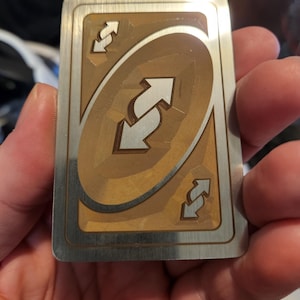 Puede incluir: Una tarjeta Uno de metal con el símbolo de inversión. La tarjeta tiene un centro dorado con un borde plateado. El símbolo de inversión es una flecha blanca dentro de un óvalo dorado, con flechas de inversión más pequeñas en cada esquina.