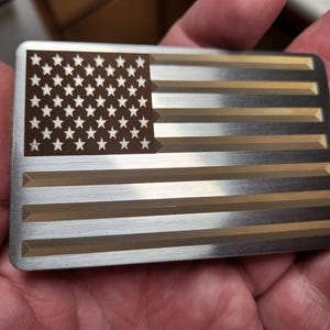 Könnte beinhalten: Ein rechteckiges Metallobjekt mit dem Design der amerikanischen Flagge. Die obere linke Ecke hat einen braunen Bereich mit weißen Sternen, der Rest abwechselnd silberne und goldene Streifen. Das Objekt wird in einer Hand gehalten.