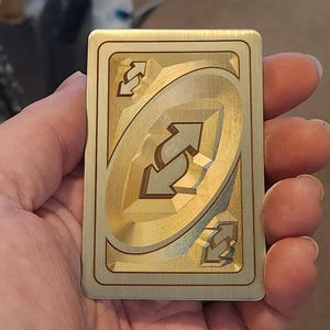 Puede incluir: Una tarjeta metálica dorada con el símbolo de inversión de dirección del juego de cartas Uno. La tarjeta tiene un acabado de metal cepillado con un diseño grabado detallado. El símbolo de inversión de dirección está en el centro.