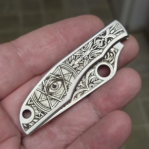 May include: A silver-toned folding pocket knife handle featuring intricate laser-engraved details. The design includes a prominent eye centered within a geometric star pattern, surrounded by elaborate floral and scrollwork motifs across the surface.