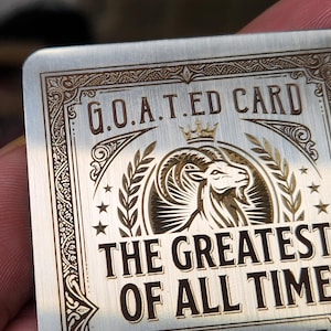 Puede incluir: Una tarjeta metálica rectangular con texto y diseño grabados. La tarjeta dice "GOATED CARD" encima de una ilustración de cabeza de cabra, y "THE GREATEST OF ALL TIME" debajo. La tarjeta tiene bordes decorativos y estrellas.