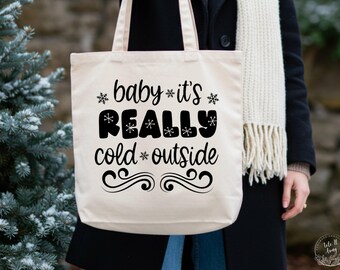 May include: A beige tote bag with the phrase "baby it's REALLY cold outside" in black script and snowflake accents. The bag has a simple design, perfect for carrying everyday items. The bag is held by a person wearing a black coat and a white scarf.