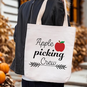 May include: A cream tote bag featuring the text "Apple Picking Crew" in black script, a red apple graphic, and an arrow design. The bag is held by a person wearing a black jacket. Pumpkins are visible in the background.