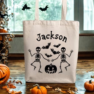 Puede incluir: Una bolsa de mano de Halloween beige con el nombre "Jackson" impreso en negro. La bolsa presenta un diseño de dos esqueletos, murciélagos y una calabaza. La bolsa está hecha de una tela resistente y tiene dos asas.