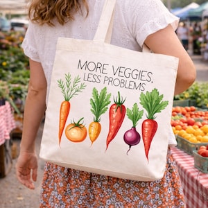 Könnte beinhalten: Beige Tragetasche mit dem Text "MORE VEGGIES, LESS PROBLEMS" und Gemüseillustrationen. Die Gemüse sind Karotten, eine Tomate und eine Radieschen. Die Tasche wird auf einem Markt getragen.