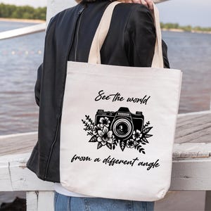 Op de afbeelding: Een witte tote bag met zwarte tekst en een zwart-witte illustratie van een camera met bloemen. De tekst luidt "See the world from a different angle."