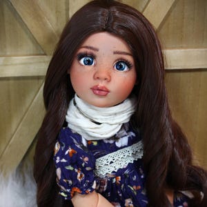 May include: A handcrafted doll with long, dark brown hair and bright blue eyes. The doll wears a navy blue floral dress with a white scarf and a red bracelet. The doll's face has freckles and a soft, rosy complexion.