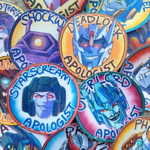 Può includere: Una collezione di adesivi rotondi e colorati con volti di robot stilizzati. Ogni adesivo presenta il volto di un personaggio, un nome e la parola "APOLOGIST" in un design circolare. Gli adesivi sono in vari colori, tra cui blu, arancione e viola.