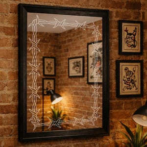 May include: A black framed mirror with a white barbed wire design, surrounded by framed tattoo art prints on a brick wall. The art includes a panther, eagle, wolf, and skull.