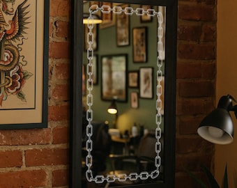 May include: A rectangular black framed mirror with a white chain design around the edges. The mirror is hanging on a red brick wall. A framed tattoo art print is visible to the left.