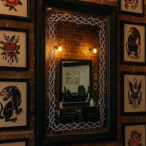 Könnte beinhalten: Ein schwarz gerahmter Spiegel mit einem weißen Dornendesign an den Rändern hängt an einer Backsteinmauer. Gerahmte Tattoo-Kunstdrucke in schwarzen Rahmen umgeben den Spiegel. Der Raum wird mit warm getönter Beleuchtung beleuchtet.