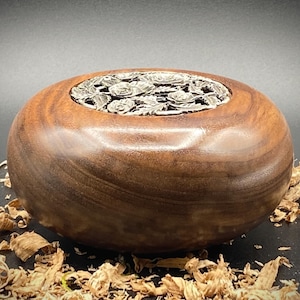 May include: A dark brown wooden bowl with a silver metal top featuring a floral design. The bowl is sitting on a bed of wood shavings.