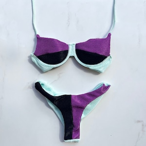 May include: A two-piece swimsuit set in a color-block design. The top is a balconette style with purple and black panels, and the bottom is a high-cut brief. The swimsuit has a textured fabric and is in shades of purple, black, and light blue.