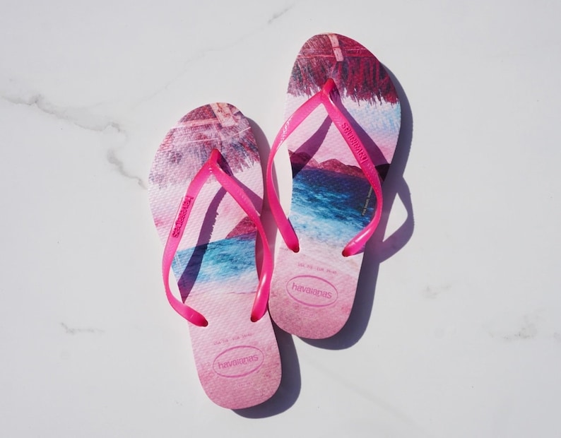 Op de afbeelding: Een paar roze slippers met een blauwe en roze strandprint. De slippers hebben een roze band en de merknaam "havaianas" staat op de zool gedrukt.